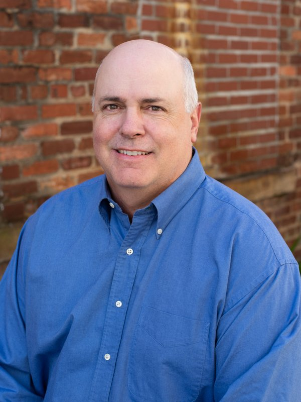Smiling bald man in blue button-down shirt standing in front of brick wall