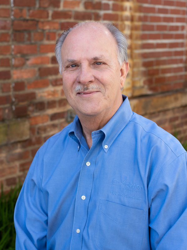 Smiling older man with grey hair and mustache wearing blue collared shirt, standing in front of brick wall.