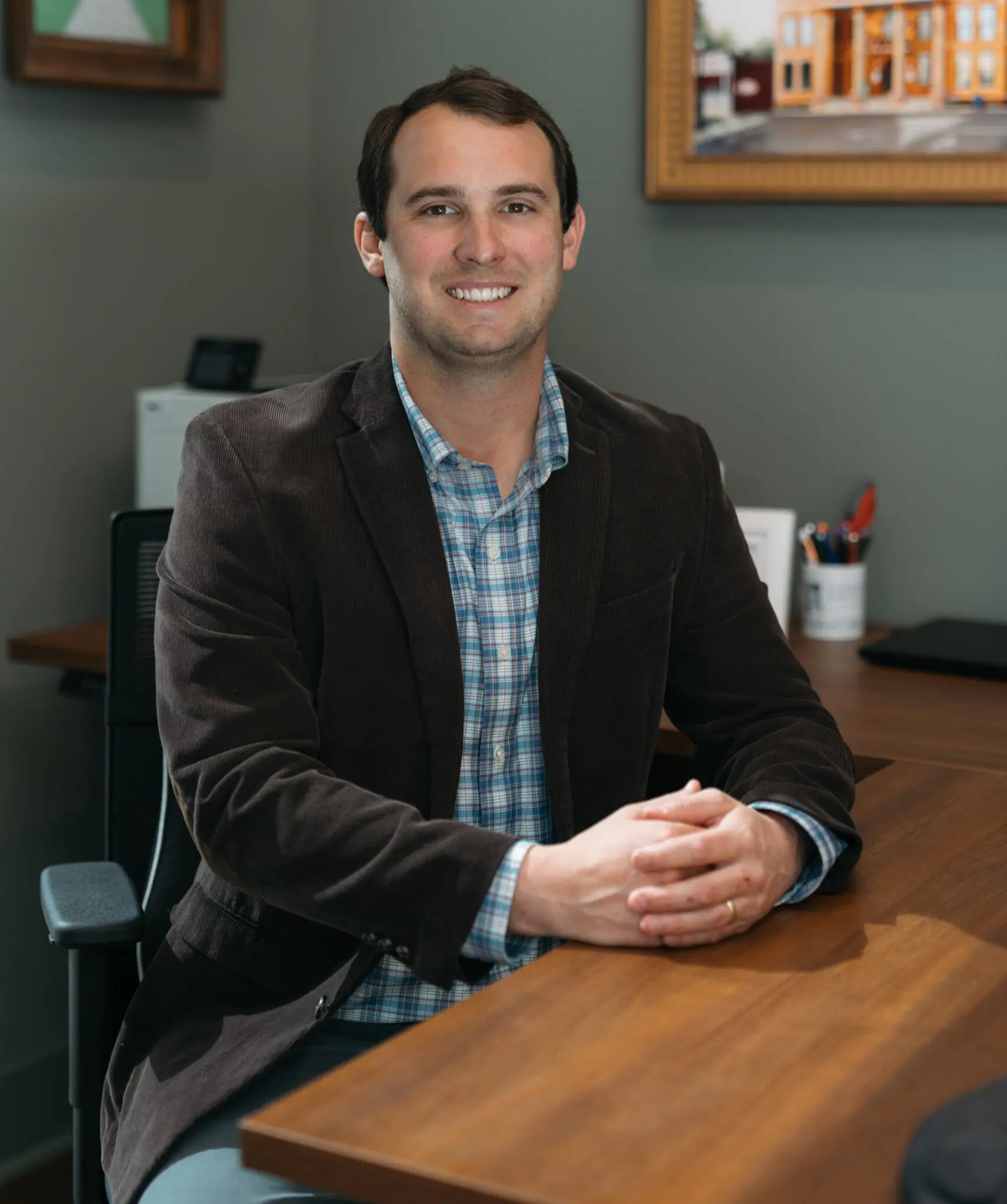 Smiling businessman in brown blazer and blue plaid shirt at office desk, friendly professional portrait