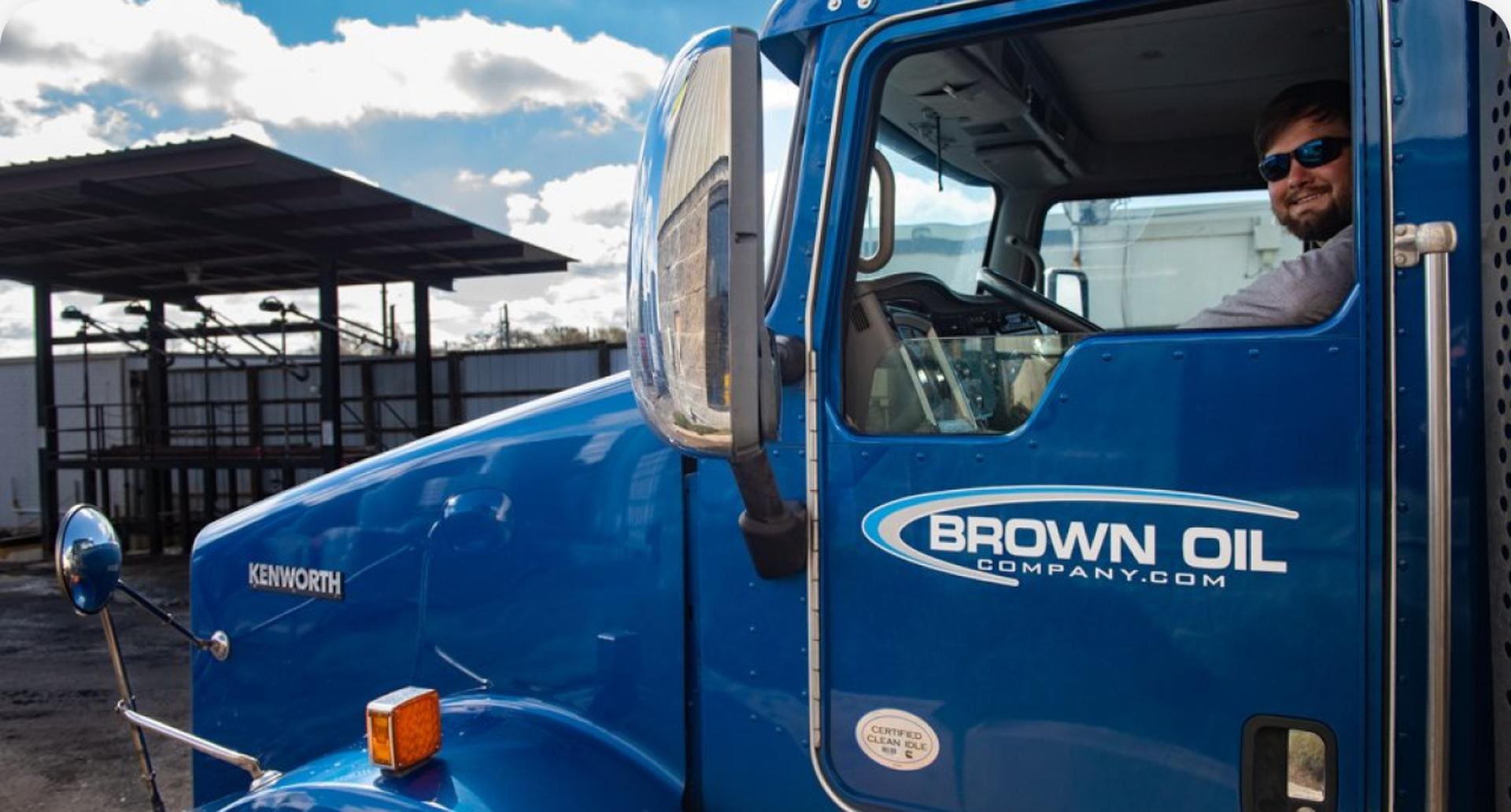 Blue Brown Oil Company truck with driver at the wheel parked under a roof structure with sunlight