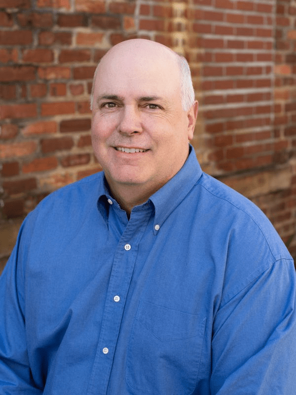 Smiling bald man in blue button-down shirt standing in front of brick wall