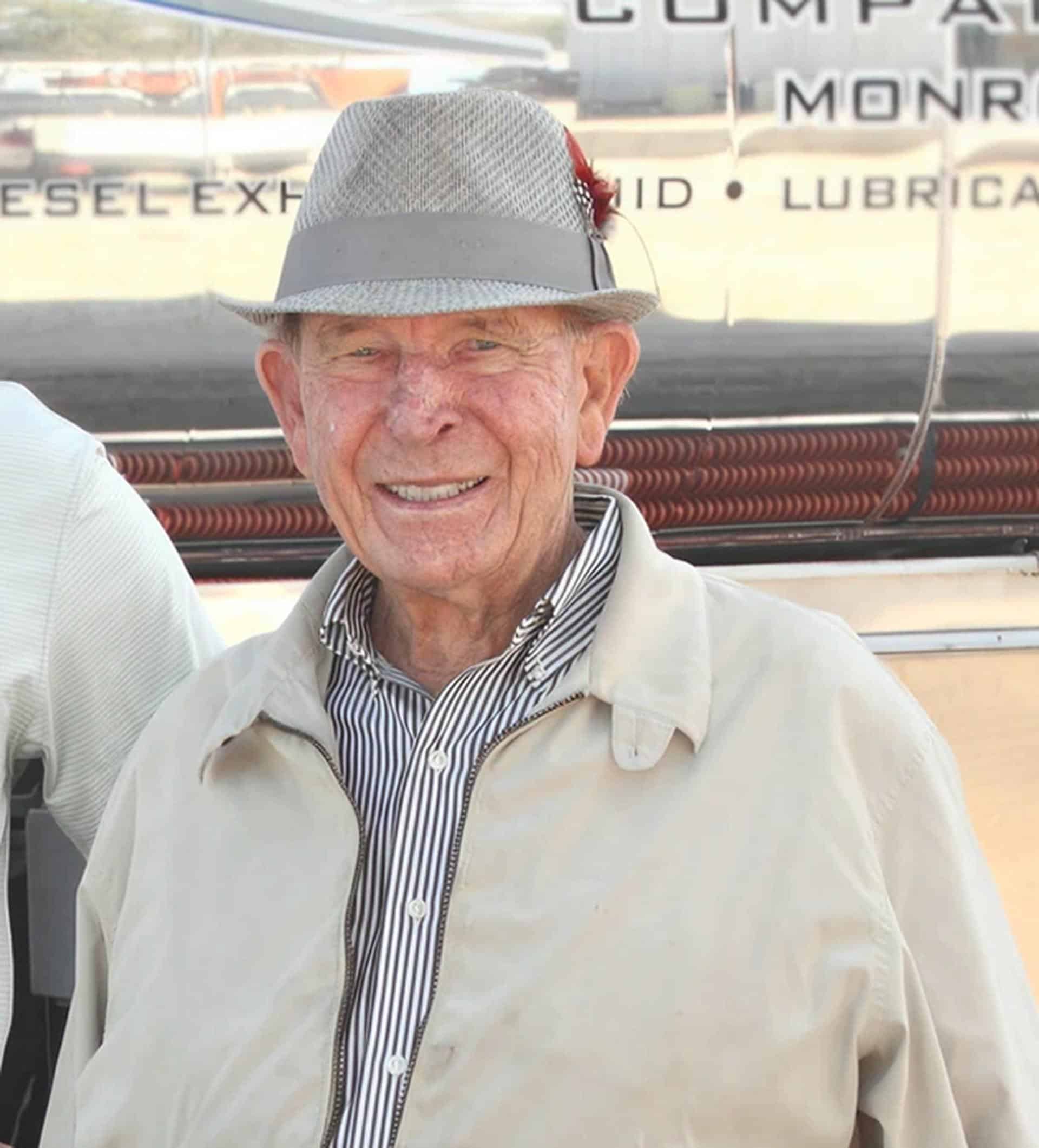 Elderly man wearing fedora smiling in front of vintage diesel truck with lubricant company sign
