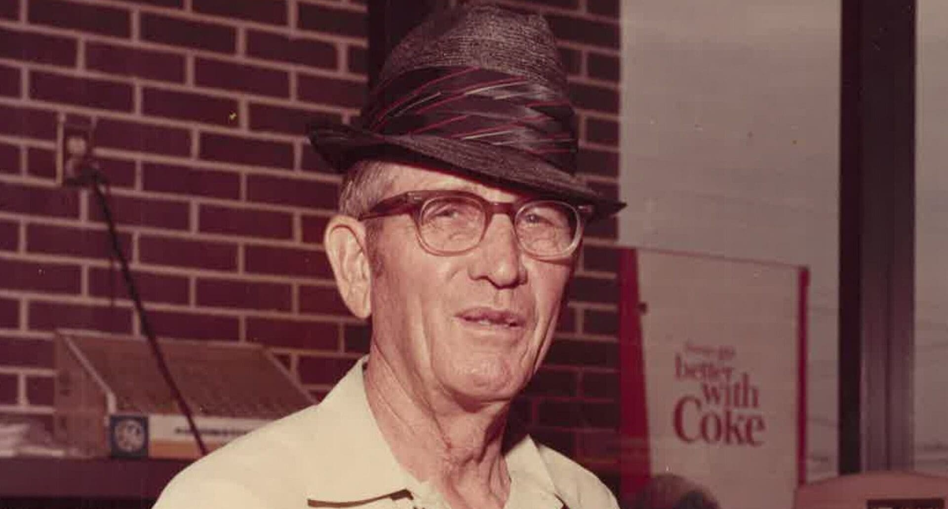 Elderly man wearing glasses and hat standing in front of brick wall with Coca-Cola sign