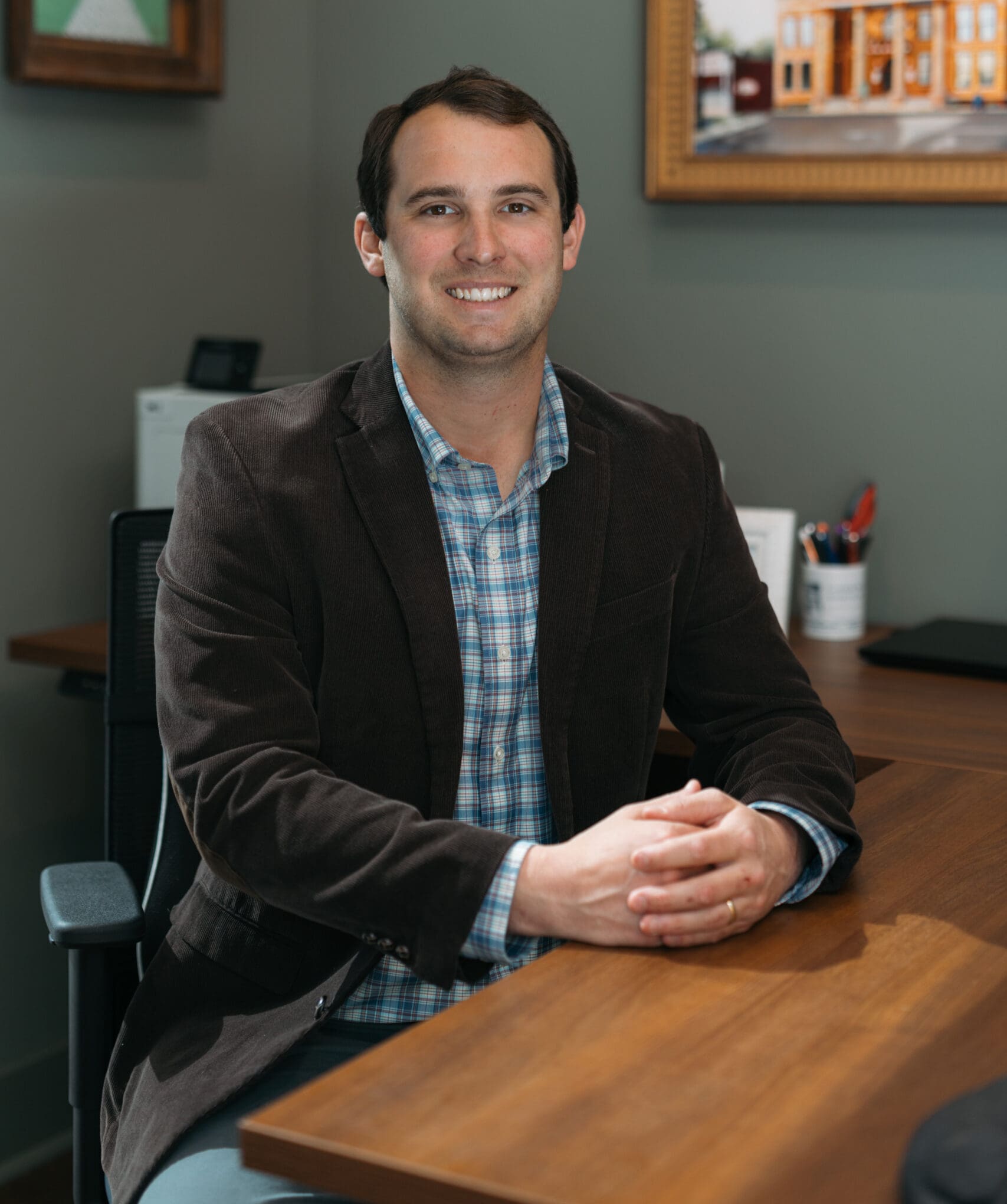 Smiling businessman in brown blazer and blue plaid shirt at office desk, friendly professional portrait